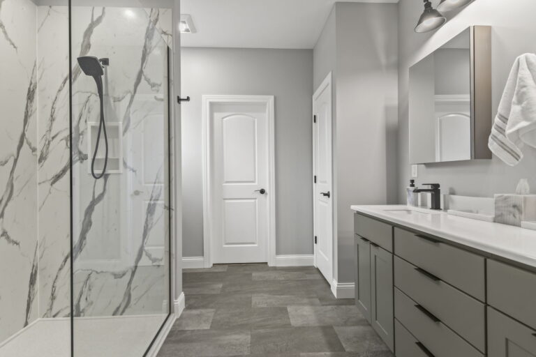 Classic modern bathroom with large walk in shower. Warm grays and whites come together with Showplace Cabinetry in Dovetail Paint, Adura LVT flooring in Steele, MSI Premium Plus White Quartz tops, and Calacatta look shower panels. Located in Centennial Park Place, Springfield, IL