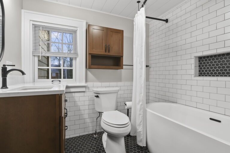 Toto toilet, overhead wooden additional storage showing both the woodedn vanity with quartz top, white soaking tub, subway tile, black hexagon tile flooring with black grout.